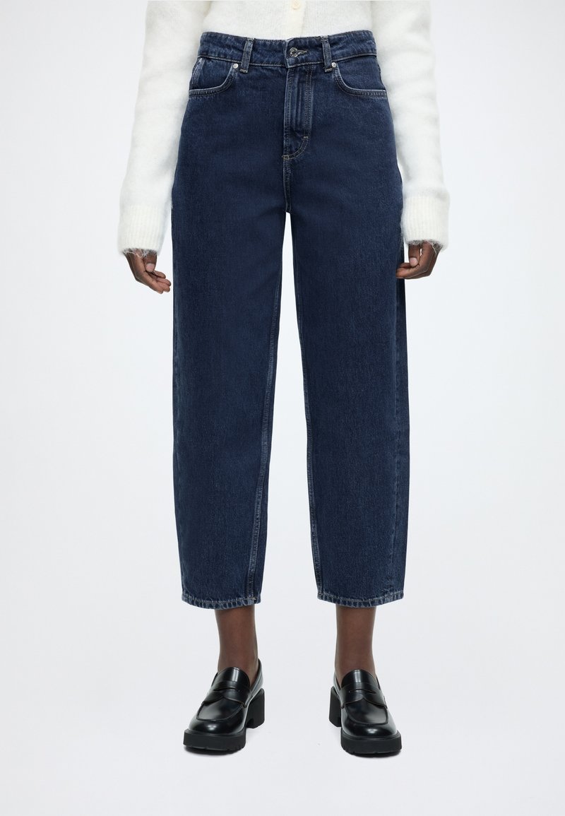 Τζιν σκούρου χρώματος, με ψηλή μέση και ίσια γραμμή, πέντε τσέπες, αντίθετη ραφή και ακατέργαστο τελείωμα. Συνδυασμένο με μαύρα μοκασίνια με πλατφόρμα.