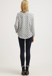 Kobieta z blond włosami nosi szary, w kratkę, długi rękaw, ciemne dopasowane dżinsy podwinięte na mankietach oraz czarne, skórzane botki na obcasie, stoi tyłem.