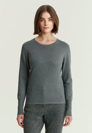 Femme portant un pull gris ajusté à manches longues et un pantalon gris texturé, debout devant un fond clair uni.