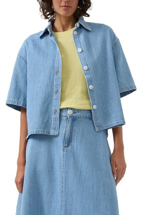 Kobieta w jasnoniebieskiej krótkiej kurtce dżinsowej z białymi guzikami, na żółtej koszulce oraz dopasowanej, wysokiej spódnicy dżinsowej.