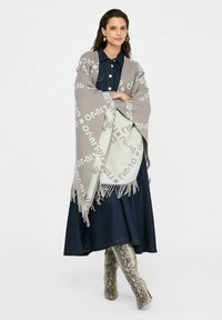 Sciarpa grigia intrecciata con frange, con motivo ripetuto in grigio più chiaro. Indossata sopra un vestito di jeans e abbinata a stivali con stampa di serpente.