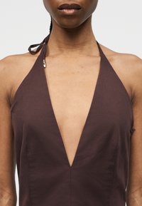 Κοντινό πλάνο ενός ατόμου που φοράει σκούρο καφέ τοπ χωρίς μανίκια, με βαθύ V ντεκολτέ και δεμένη λουρίδα στο λαιμό.