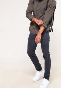 Camicia olive green con maniche arrotolate, abbinata a jeans slim-fit grigio scuro e sneakers bianche, con uno zaino nero.
