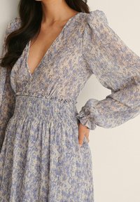 Šaty s květinovým vzorem v odstínech modré a broskvové, s hlubokým V výstřihem, dlouhými puff rukávy, smockovaným pasem a volánkovými manžetami.
