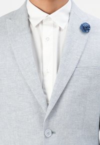 Ανοιχτό γκρι σακάκι με μπλε υφασμάτινη καρφίτσα πέτου, φορεμένο πάνω από ένα λευκό πουκάμισο με κουμπιά και μυτερό γιακά.