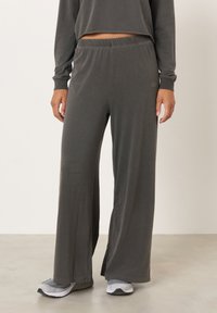 Asmuo dėvi laisvas tamsiai pilkas kelnes ir atitinkantį viršutinį drabužį su pilkais ir baltais sportbačiais, stovi ant šviesios grindų dangos priešais paprastą foną.