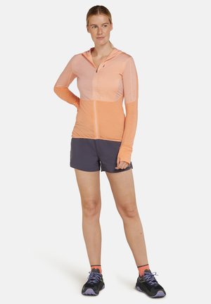 Lätt zip-up hoodie i orange nyanser med genomskinliga paneler, kombinerad med mörka shorts. Har en huva och tumhål i ärmarna för bättre skydd.
