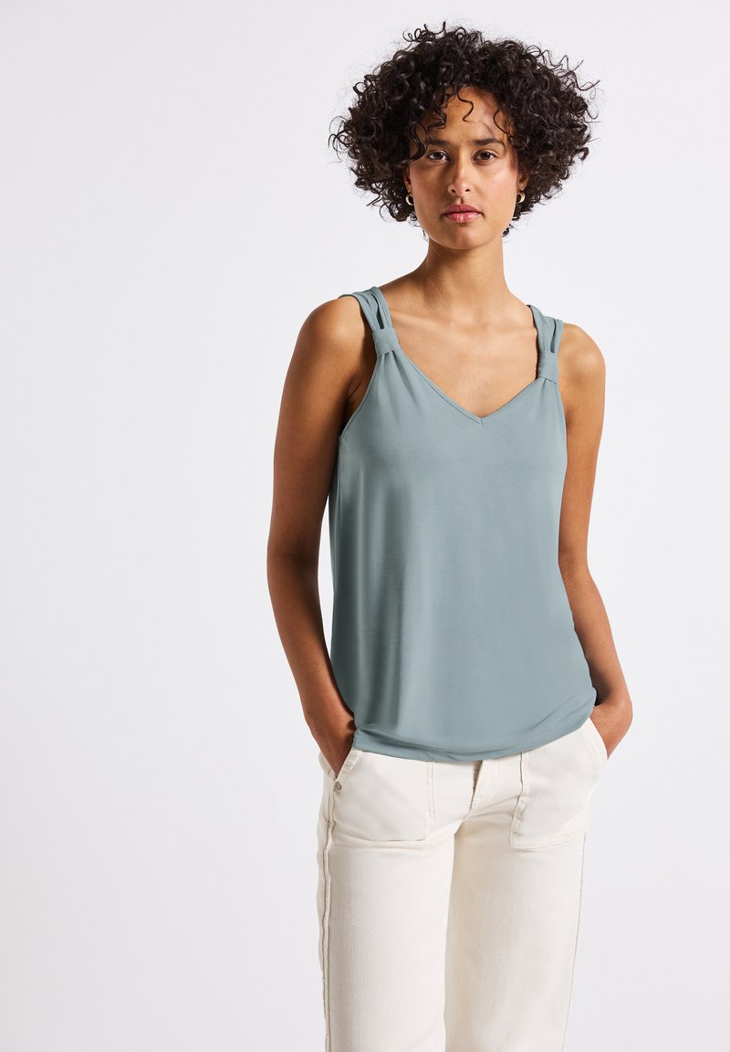 Ljusblå ärmlös topp med V-ringning och dekorerade remmar. Tillverkad av ett glatt tyg; kombinerad med vita, avslappnade byxor.