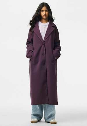 Jeune femme aux longs cheveux foncés portant un long manteau violet, un pull violet clair, un jean large bleu et des baskets blanches, debout les mains dans les poches.