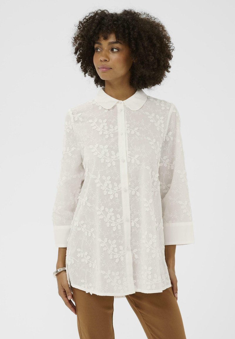 Žena s kučeravými vlasmi, oblečená v priesvitnej bielej výšivkovej blúzke na gombíky a v hnedých nohaviciach, pozerá sa bokom na jednoduchom pozadí.