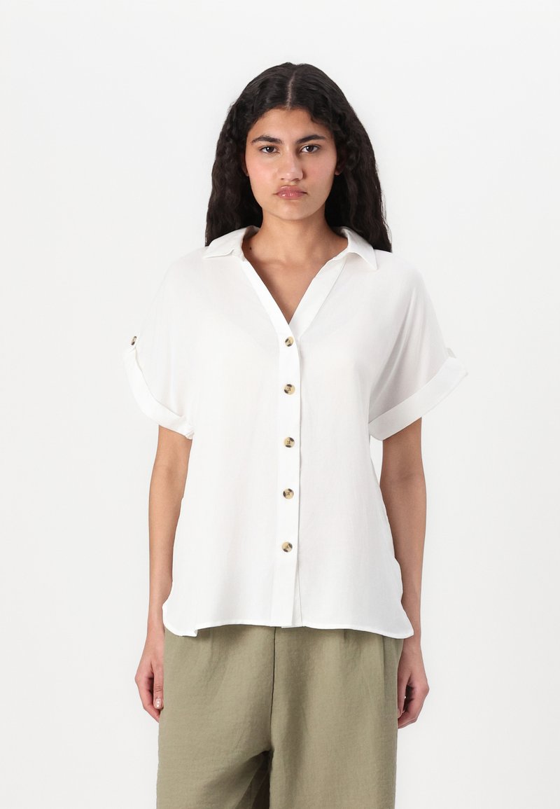 Blusa bianca a maniche corte con chiusura a bottoni sul davanti, leggero lucido, e maniche arrotolate; il tessuto sembra leggero e liscio.