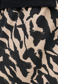 Ύφασμα με αφηρημένο ζωικό μοτίβο σε μαύρο και μπεζ χρώμα, με ακανόνιστες, σαν πινελιές μορφές, κάτω από ένα στερεό μαύρο ένδυμα.