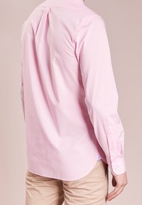 Ljusrosa långärmad skjorta med knappdetaljer, enkelstygnad krage och rundad fåll. Slät bomullstyg. Synlig veck bak.