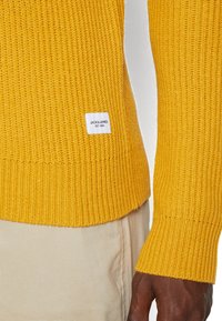 Gelber Strickpullover mit Rippenstruktur und Bündchen, an der Saumkante mit einem kleinen weißen Etikett "JACK & JONES EST. 1990".
