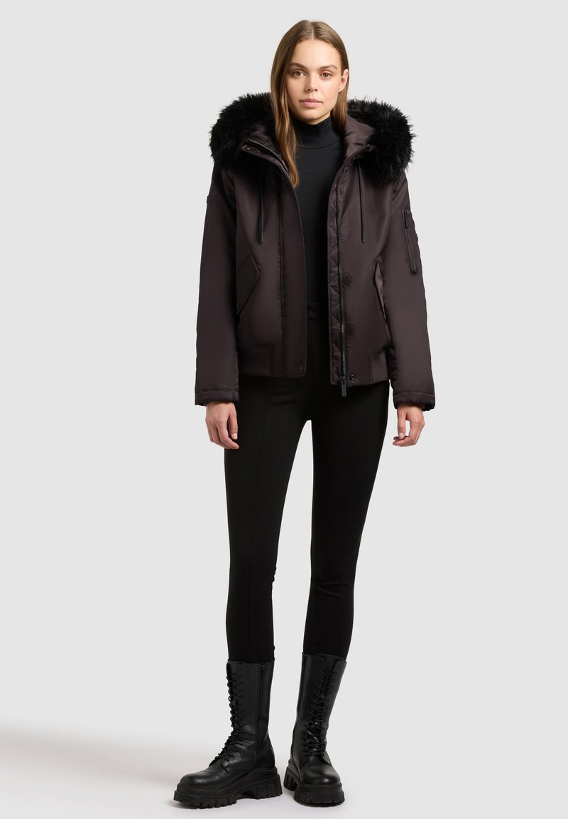 Schwarze Daunenjacke mit einer Kapuze aus Kunstfell, Reißverschluss- und Knopfveschluss sowie seitlichen Taschen. Tragen über einem schwarzen Rollkragenpullover und Leggings.