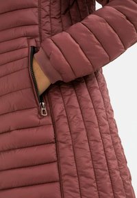 Quilted padded jacket in dusty rose with a zippered pocket and ribbed cuffs. Features a smooth texture and stitched horizontal patterns.