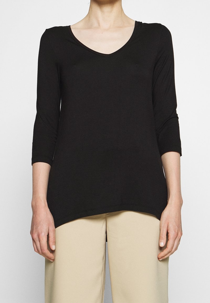 Nainen, jolla on päällään musta V-kaula-aukko ja kolme neljännesosaa hihasta oleva paita sekä vaaleat housut, seisoo tasaista vaaleaa taustaa vasten.