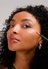 Femme aux cheveux bouclés applique un correcteur avec un pinceau sur sa joue près de l'œil, portant des boucles d'oreilles en or et un rouge à lèvres brillant.