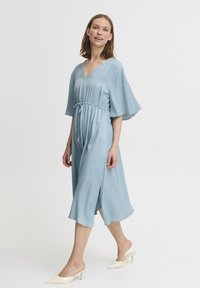 Hellblaues Midi-Kleid mit V-Ausschnitt, tailliertem Bund, fließenden Ärmeln und seitlichen Schlitzen, kombiniert mit beigen Absatzsandalen.