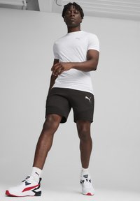 Camiseta blanca ajustada, pantalones cortos negros con el logo de Puma y zapatillas deportivas con acentos rojos y negros. Tejido suave y diseño casual.