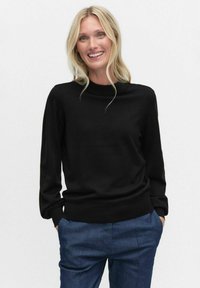 Mustan neulotun neuleen korkea kaula ja pitkät hihat. Sujuva rakenne, vartalonmyötäinen malli vyötäröllä. Yhdistetty sinisiin housuihin.