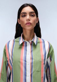 Camicia button-up a righe nei colori verde, rosa e bianco, con linee orizzontali di larghezze variabili e una tasca sul petto ricamata.