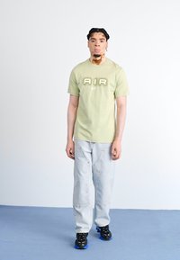 Πράσινο βαμβακερό T-shirt με την ένδειξη "AIR" σε λευκό χρώμα και το λογότυπο της Nike, συνδυασμένο με ανοιχτό μπλε τζιν παντελόνι και μαύρα sneakers με μπλε λεπτομέρειες.