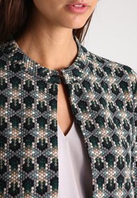 Textured, patterned jacket featuring a geometric design in dark green, beige, and black, with a round collar and buttonless front closure.