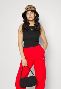 Giovane donna che indossa un cappello a secchiello con stampa leopardata, top senza maniche Adidas nero, pantaloni Adidas rossi, e tiene in mano una borsa nera.