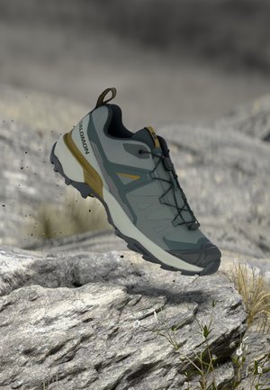 Grauer und gelber Salomon Trailrunning-Schuh schwebt über felsigem Gelände mit Erdpartikeln und kleinen Pflanzen in der Nähe.