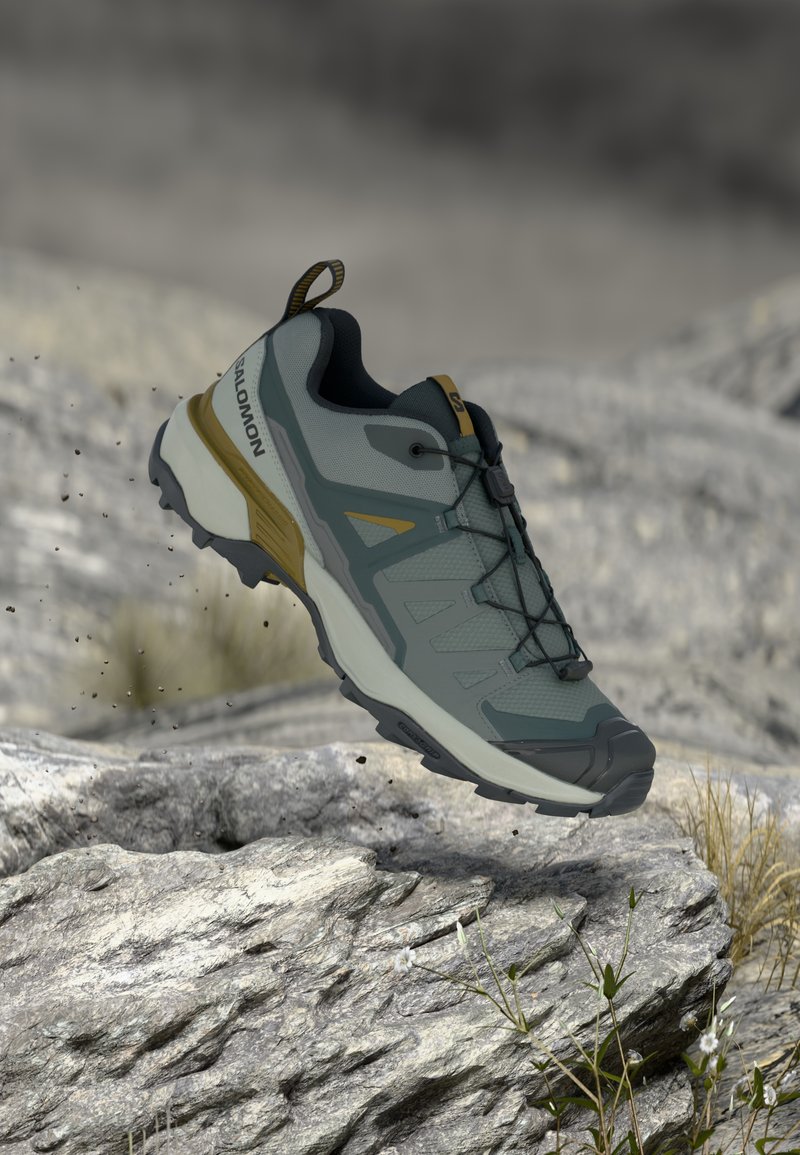 Γκρι και κίτρινο παπούτσι trail running Salomon κρεμασμένο πάνω από βραχώδες έδαφος με σωματίδια βρωμιάς και μικρά φυτά κοντά.