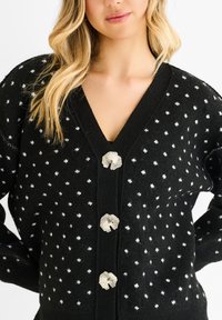 Black cardigan with white polka dots, V-neck design, and three decorative shell-shaped buttons at the front. Soft, textured fabric.