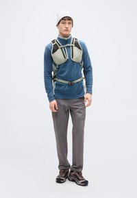 Ελαφρύ γκρι γιλέκο πεζοπορίας με ρυθμιζόμενους ιμάντες πάνω από μπλε μπλούζα με μακρύ μανίκι, σε συνδυασμό με γκρι παντελόνια και σκούρα παπούτσια πεζοπορίας.