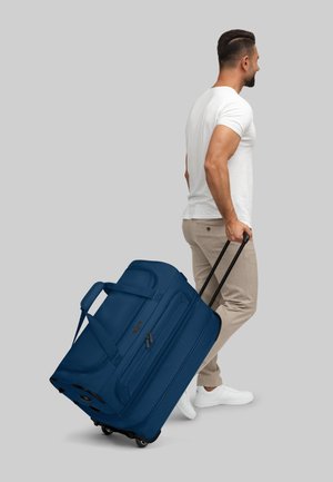 Blaue Rollreisetasche mit Reißverschlüssen und gepolsterten Griffen; strapazierfähiger Stoff und strukturierte Oberfläche; wird von einer Person in lässiger Kleidung gezogen.