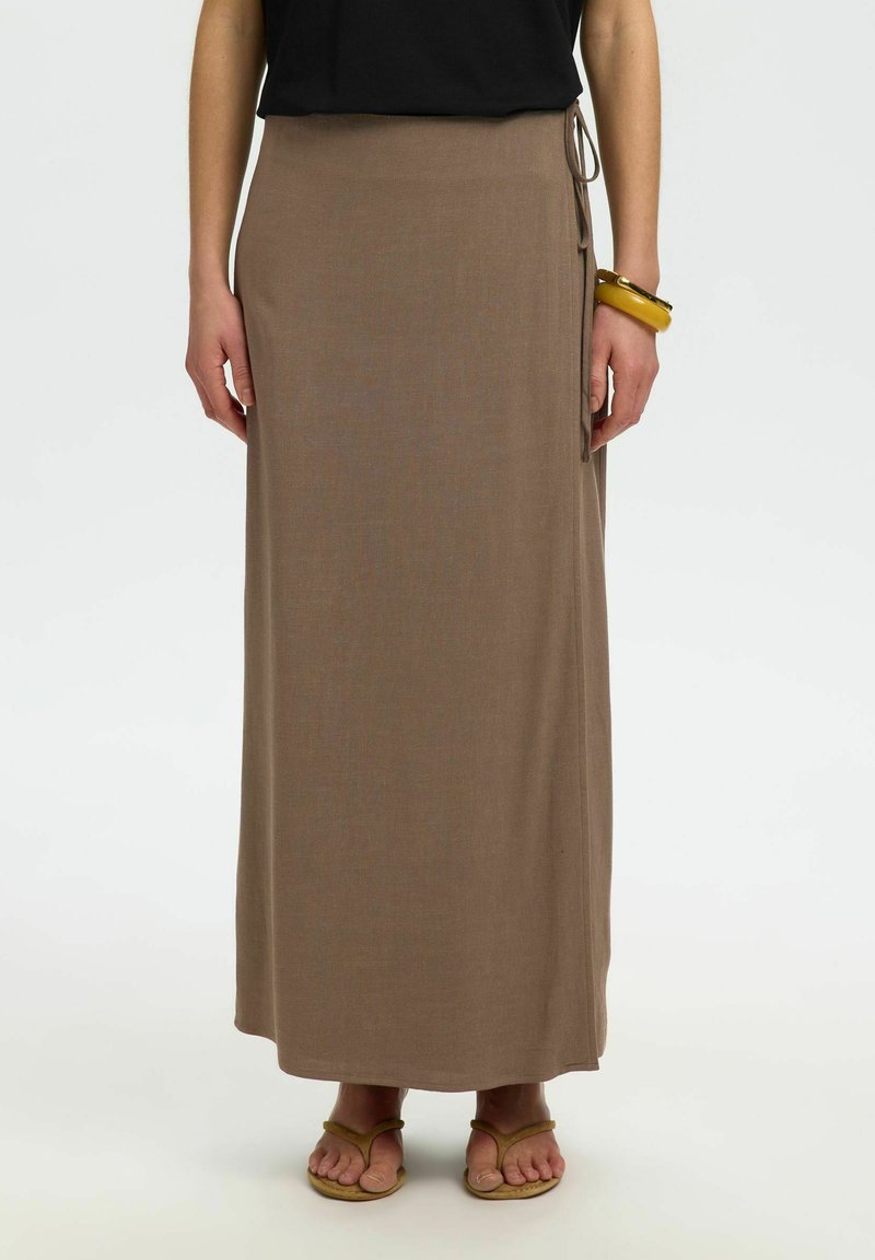 Femme portant une jupe maxi marron style portefeuille avec des liens sur le côté, associée à un haut noir, un bracelet jaune et des sandales marron.