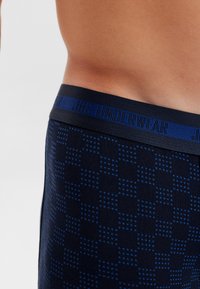 Námořnické boxerky s vzorovanou texturou, která obsahuje modré tečky a čtverce. Elastický pas zobrazuje "JBS UNDERWEAR" v tmavě modré barvě.