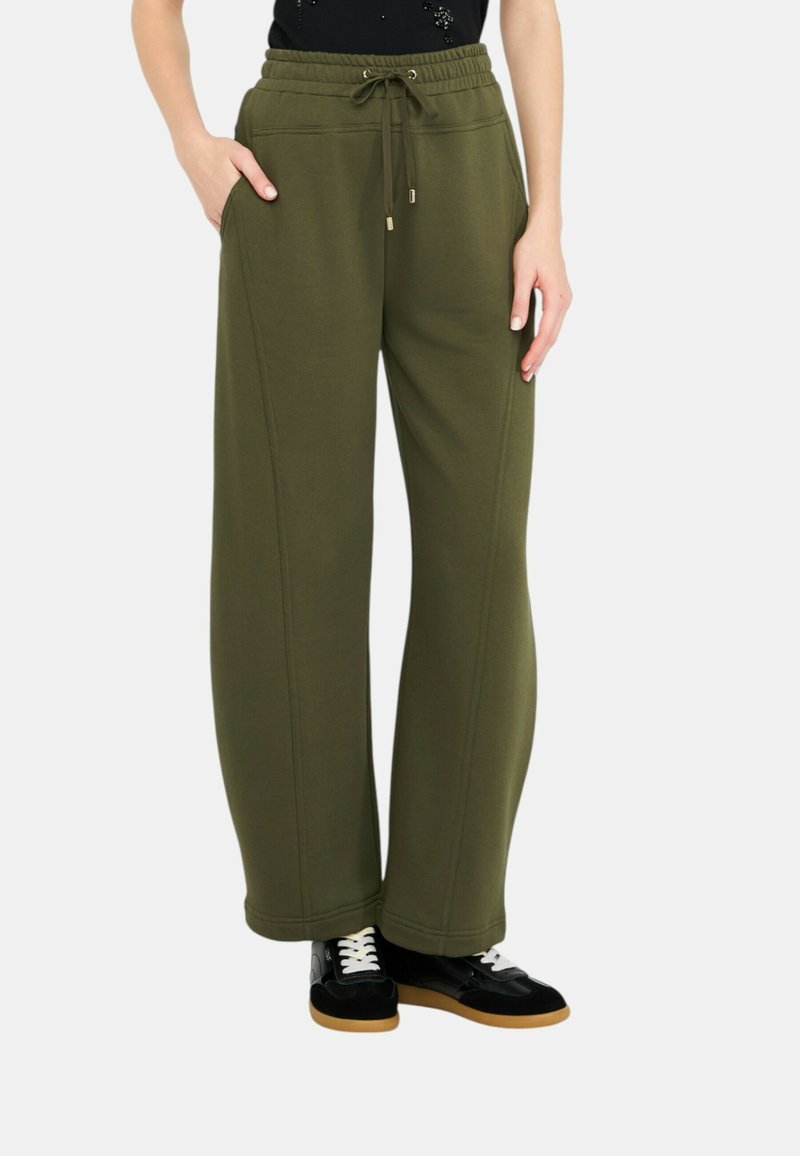 Pantaloni della tuta a gamba larga verde oliva con elastico in vita, tasche e dettagli di cucitura in vista. Abbinati a scarpe da ginnastica nere.
