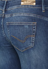 Jeans de denim azul claro com um acabamento desbotado, apresentando um pequeno bolso traseiro com costura laranja e um rótulo emblema dourado na cintura.