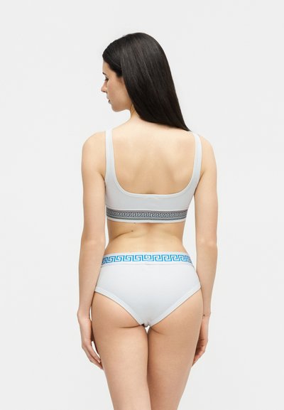 Γυναίκα με μακριά σκούρα μαλλιά που φαίνεται από πίσω, φοράει λευκό αθλητικό σουτιέν με μαύρη ταινία και λευκό εσώρουχο με μπλε ζώνη με σχέδια.