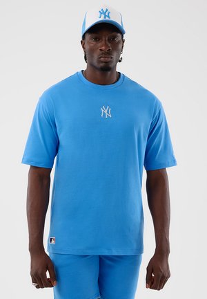 Uomo che indossa un cappellino blu dei New York Yankees, maglietta blu con logo sul petto e pantaloncini blu abbinati su uno sfondo semplice.