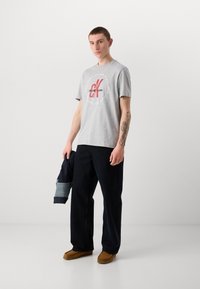 Gray cotton t-shirt with red logo, paired with dark denim pants and brown moccasins. Model holding a dark jacket with denim accents.