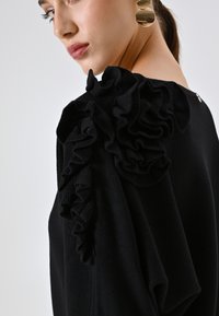 Top in maglia nera con texture arricciata sulla spalla, scollatura rotonda e tessuto morbido. Orecchini circolari dorati completano il look.