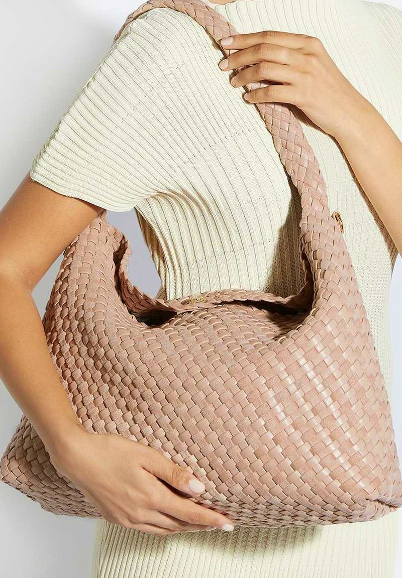 Femme portant un haut court à manches courtes côtelé crème tient un grand sac tissé rose clair avec une large bandoulière sur la poitrine.