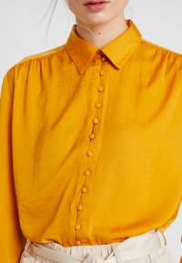 Žlutá košile na knoflíky s hladkou texturou, s klasickým límcem a knoflíky potaženými stejnou látkou vpředu.