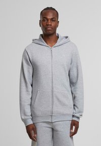 Haut à zip gris en tissu doux et brossé. Dispose d'une poche avant, d'une capuche ajustable et de poignets ajustés. S'associe avec un pantalon de survêtement gris.