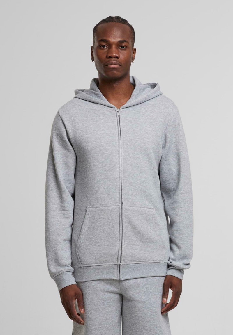 Haut à zip gris en tissu doux et brossé. Dispose d'une poche avant, d'une capuche ajustable et de poignets ajustés. S'associe avec un pantalon de survêtement gris.