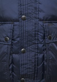 Marineblå pufferjakke med quilted tekstur, med sorte trykknapper, lynlåslukning foran og synlige logo-detaljer.