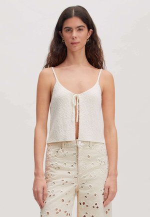 Vrouw met lang donker haar draagt een mouwloze witte gestructureerde cropped top met strik aan de voorkant en een hooggetailleerde crème broderie anglaise broek, neutrale achtergrond.