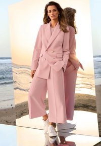 Lichtroze op maat gemaakt pak met een dubbelgeknopte blazer en wijde broekspijpen, gecombineerd met witte sneakers. Soepele stof, strakke lijnen en een moderne pasvorm.