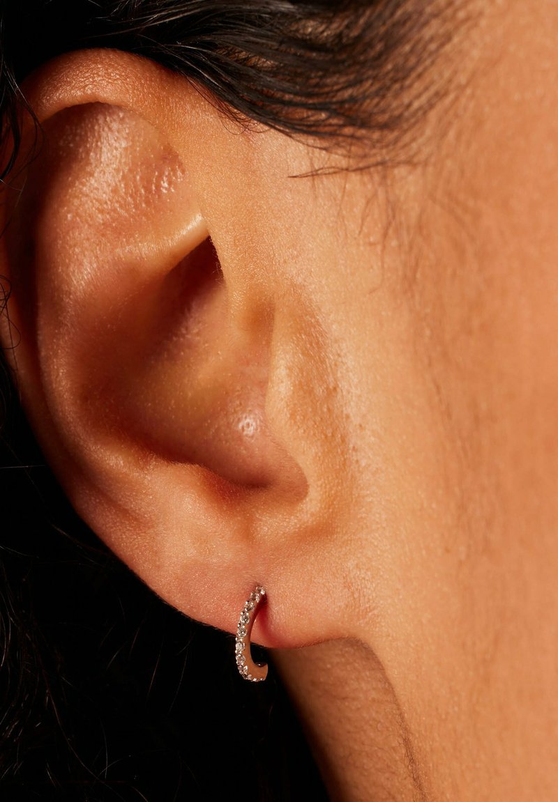 Petite boucle d'oreille en métal argenté, ornée de petits strass transparents, portée sur une oreille percée, mettant en valeur un design courbé et délicat.